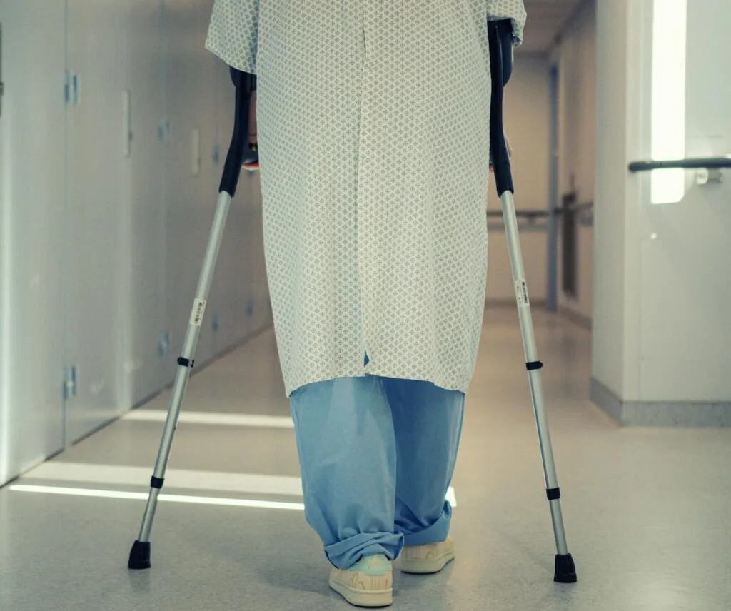 Hospital patient walking with crutches in a corridor, wearing a medical gown and blue pants