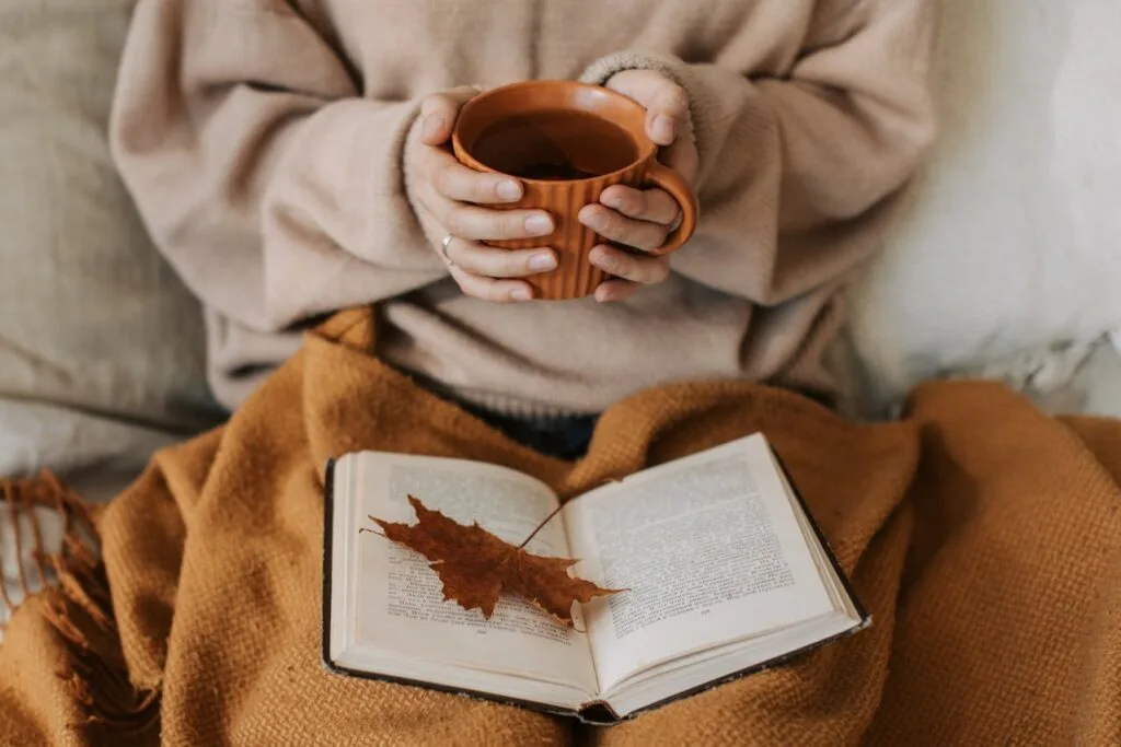 A cozy autumn scene featuring a person wrapped in a brown blanket, holding a warm mug over an open book with a dried leaf on top &mdash; perfect example of affordable autumn home decor.