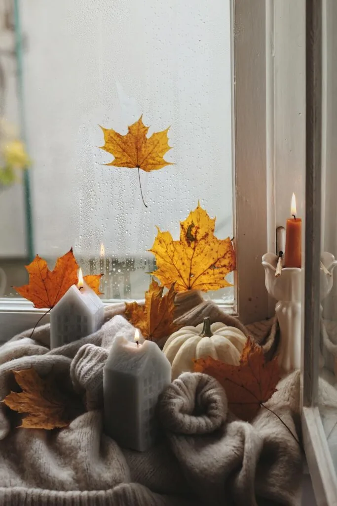 Candles, sweaters, a white pumpkin, and orange maple leaves arranged on a windowsill create a warm and inviting fall atmosphere on a rainy day.