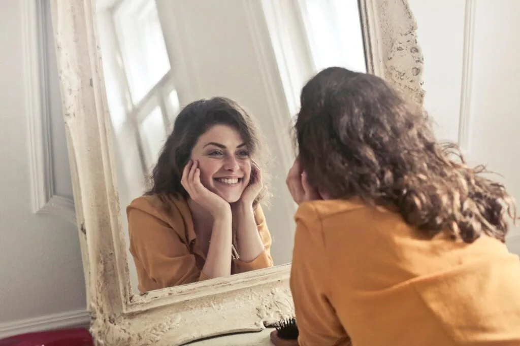 Woman smiling at her reflection in the mirror, representing self-confidence and emotional healing during balanced recovery after injury.