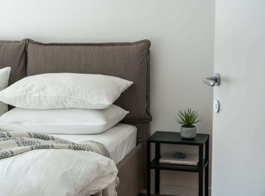 Modern bedroom with neatly stacked ergonomic pillow set on a bed beside a nightstand and plant for a clean, cozy look.