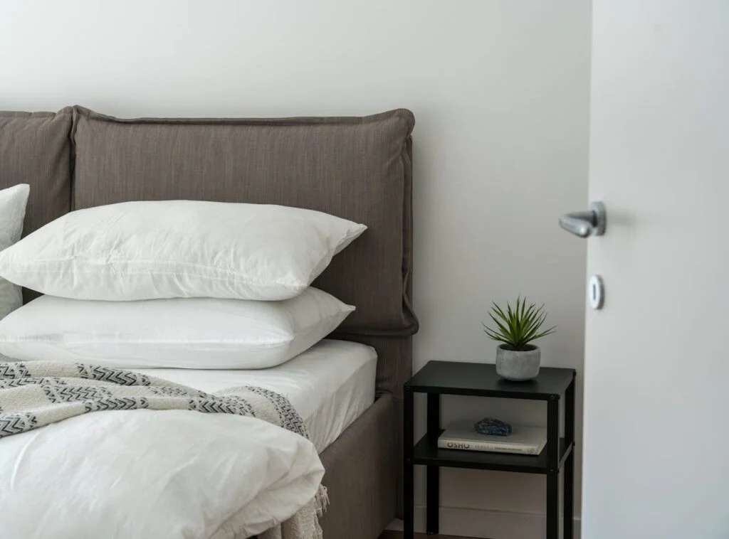 Modern bedroom with neatly stacked ergonomic pillow set on a bed beside a nightstand and plant for a clean, cozy look.