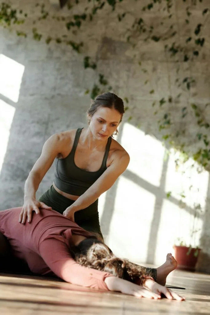 A fitness instructor assisting a client in a yoga stretch during a personal training session inside a bright, plant-filled studio.
