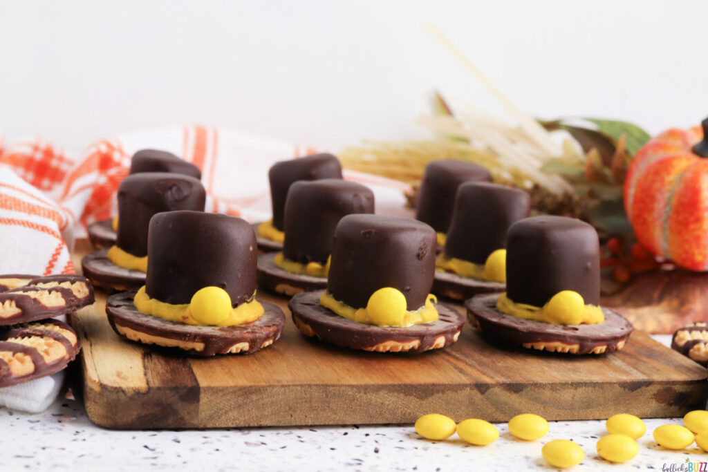Fudge Stripe Pilgrim Hat Cookies arranged on a wooden board, each topped with a chocolate-covered marshmallow and yellow candy buckle, an easy no-bake Thanksgiving dessert from Bullock’s Buzz.