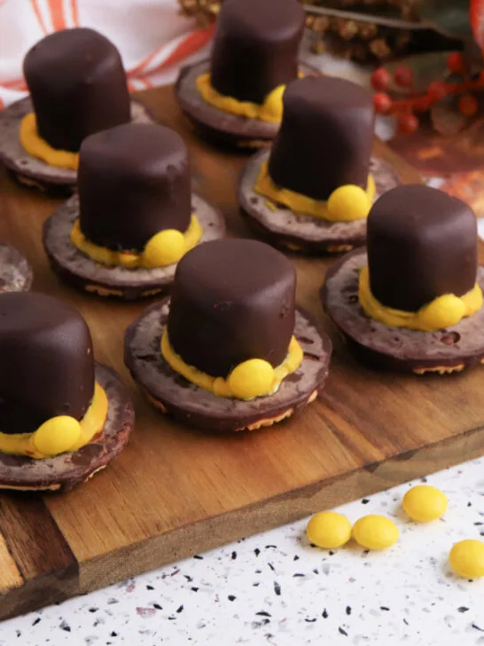 Easy no-bake Fudge Stripe Pilgrim Hat Cookies topped with chocolate-dipped marshmallows and yellow candy decorations, a festive Thanksgiving dessert idea from Bullock’s Buzz