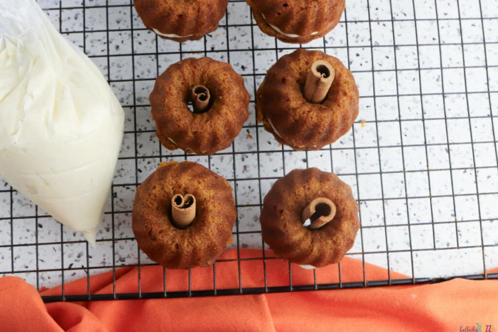 Assembling pumpkin cakes by stacking two Bundt cakes with frosting, then inserting cinnamon sticks for stems