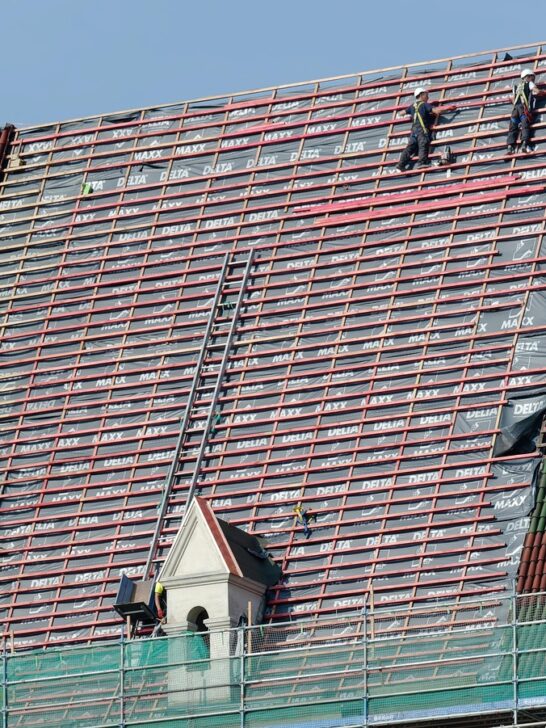 Construction workers performing roof maintenance on a large tiled roof, reinforcing beams and preparing for new shingles.