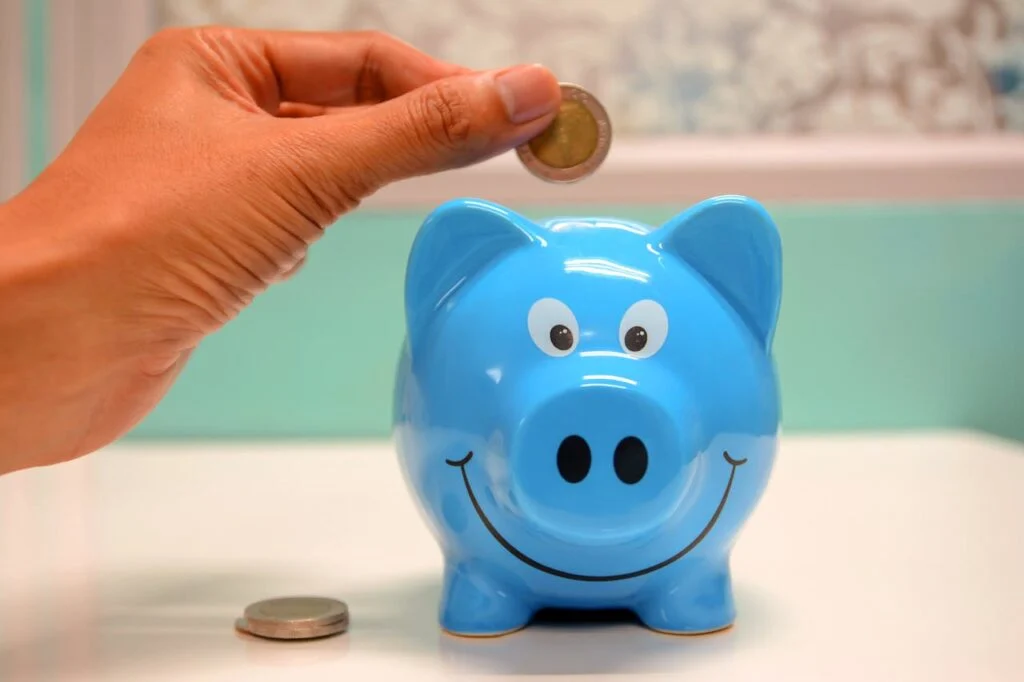 Hand placing a coin into a smiling blue piggy bank, symbolizing saving for your child&rsquo;s future.