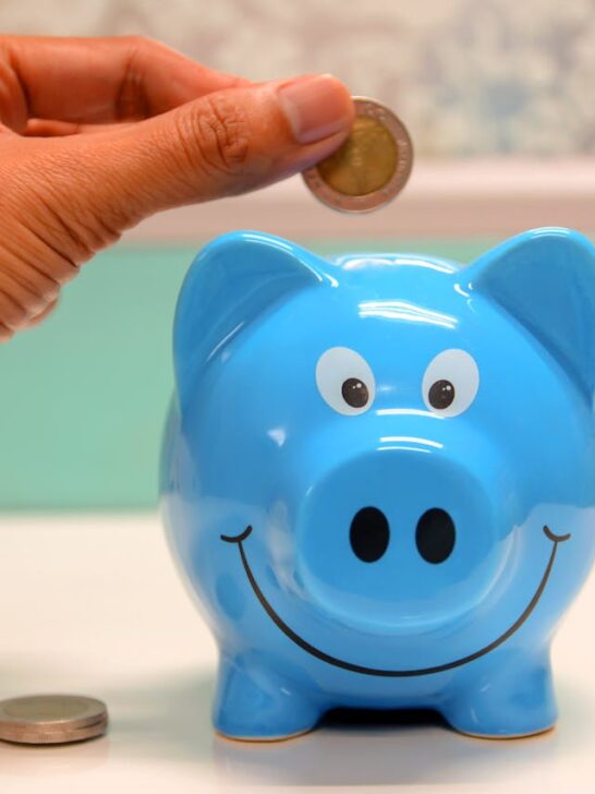 Hand placing a coin into a smiling blue piggy bank, symbolizing saving for your child’s future.