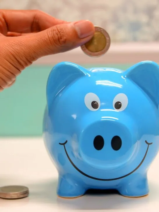 Hand placing a coin into a smiling blue piggy bank, symbolizing saving for your child’s future.
