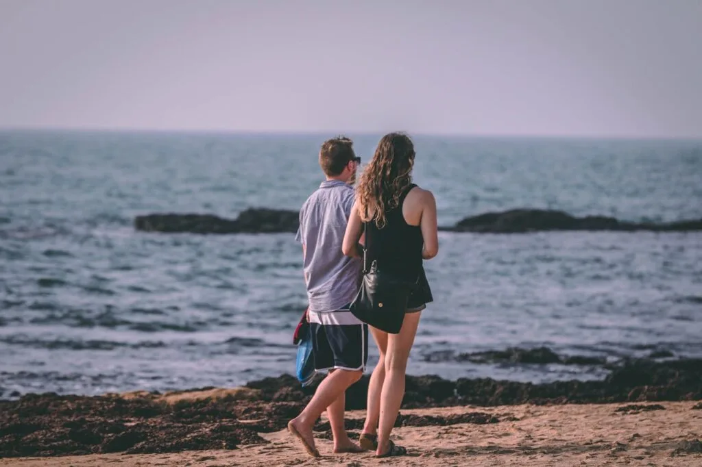 A couple walks barefoot along the beach shoreline wearing relaxed summer sun protection outfits, enjoying the ocean view under soft daylight.