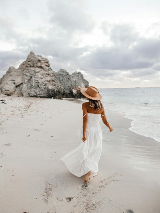 A woman in a white flowy dress and wide-brimmed hat walks along a sandy beach, showing effortless summer sun protection style against the coastal breeze.