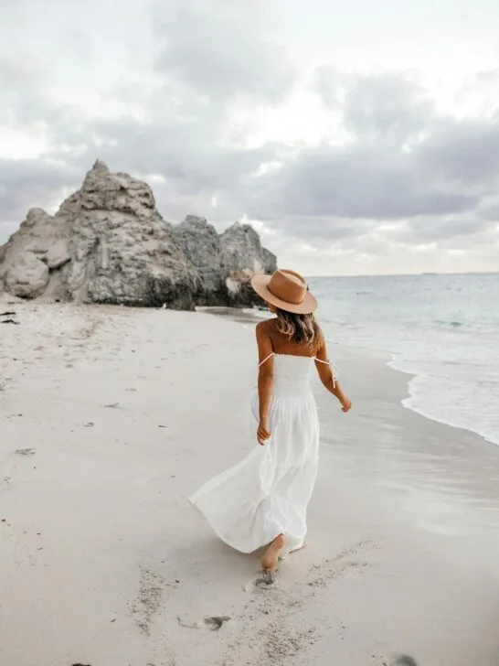 A woman in a white flowy dress and wide-brimmed hat walks along a sandy beach, showing effortless summer sun protection style against the coastal breeze.