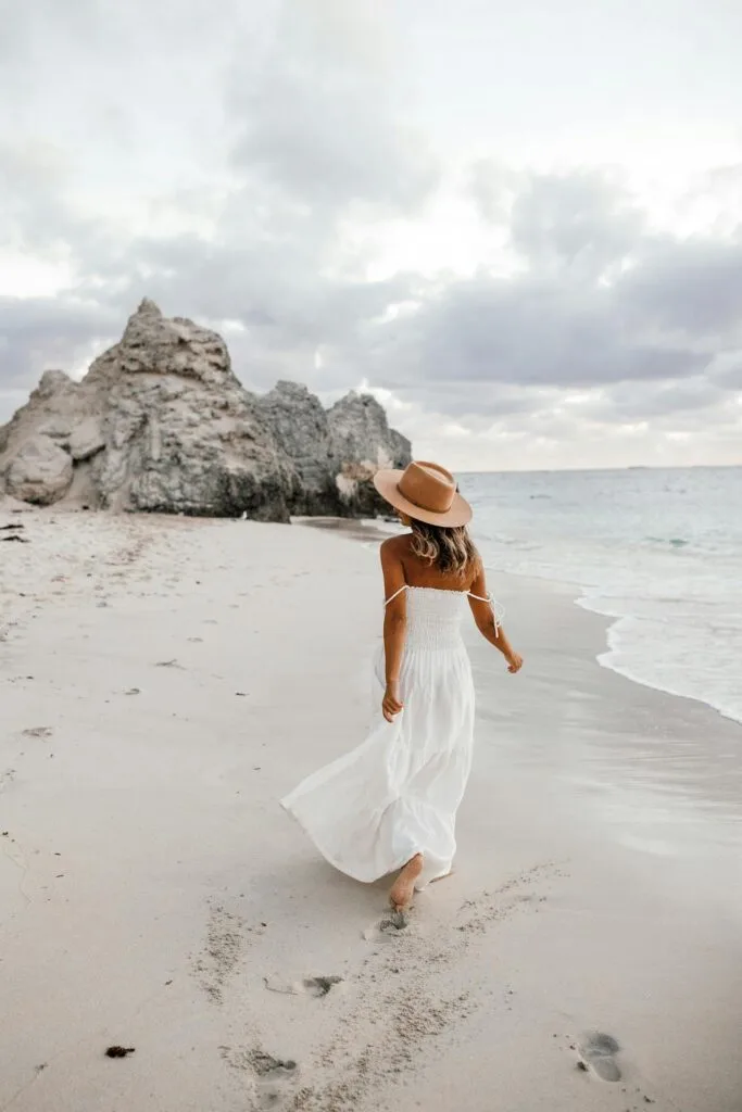 A woman in a white flowy dress and wide-brimmed hat walks along a sandy beach, showing effortless summer sun protection style against the coastal breeze.