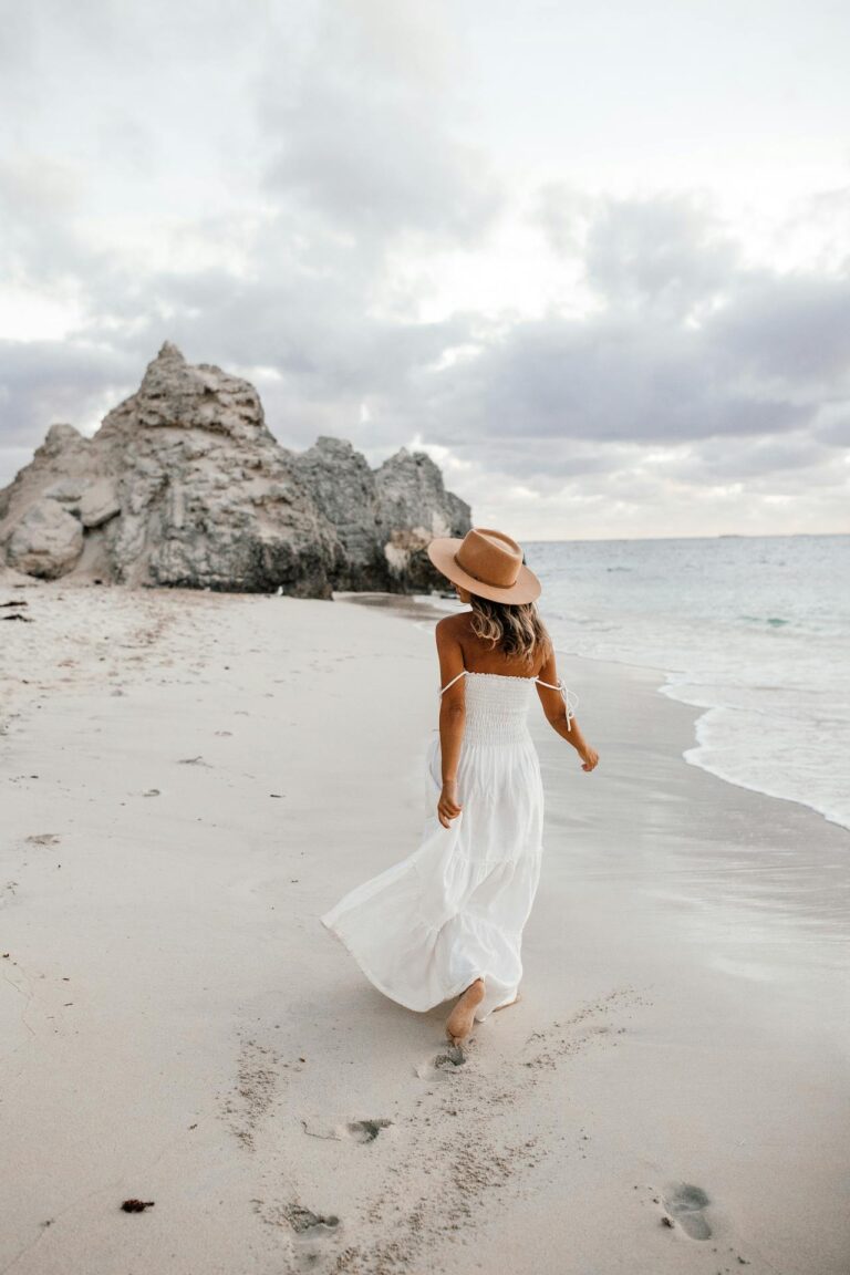 A woman in a white flowy dress and wide-brimmed hat walks along a sandy beach, showing effortless summer sun protection style against the coastal breeze.