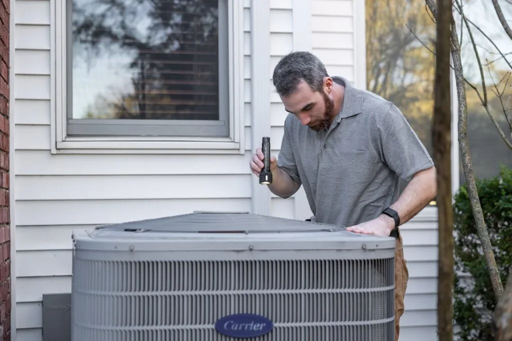 HVAC technician inspecting an outdoor air conditioning unit beside a home during a maintenance check.