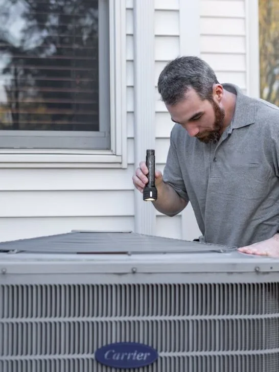 HVAC technician inspecting an outdoor air conditioning unit beside a home during a maintenance check.