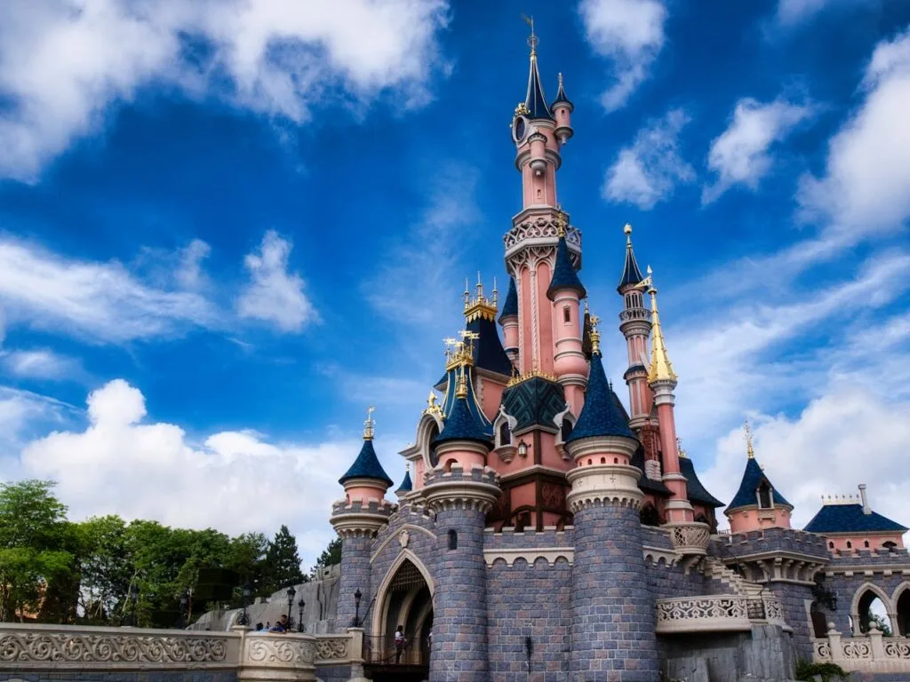 Pink and blue fairytale-style Disney castle under a bright blue sky at a theme park, surrounded by stone walls and spires.
