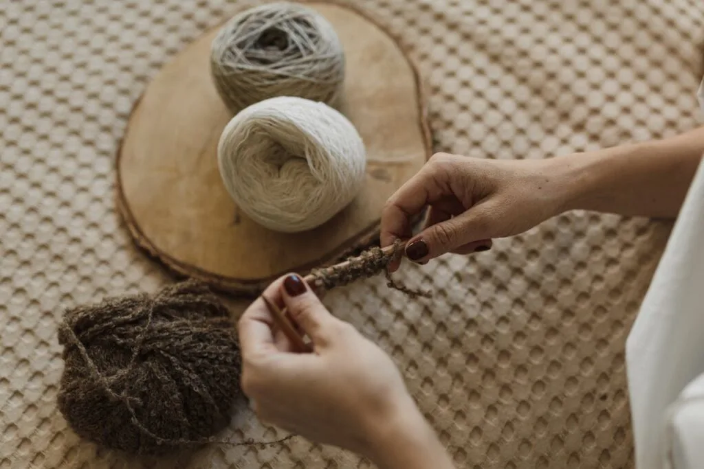 Hands holding knitting needles while working with brown yarn, with additional yarn balls resting on a wooden board.
