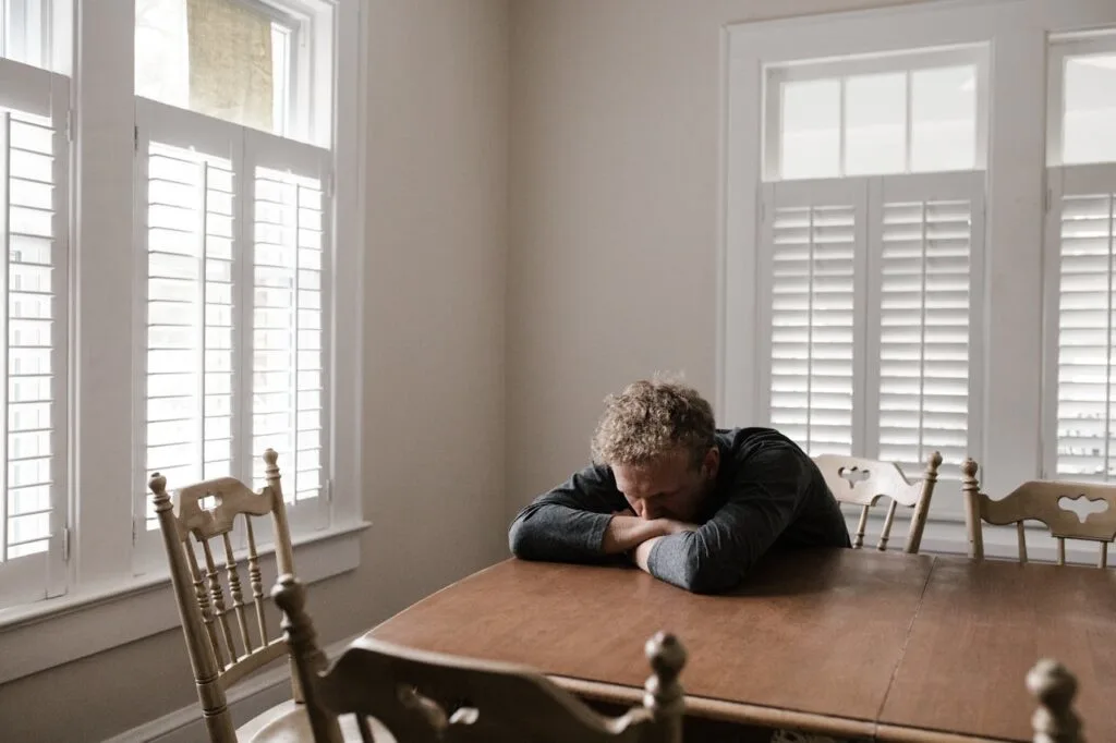 Man sitting alone with his head resting on folded arms at a wooden table, showing signs of emotional exhaustion or depression.