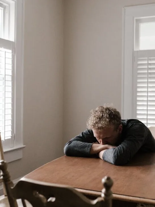 Man sitting alone with his head resting on folded arms at a wooden table, showing signs of emotional exhaustion or depression.