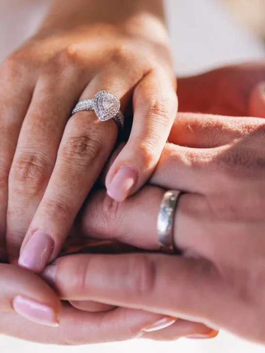 Close-up of a couple’s hands showing a diamond engagement ring and a man wearing a platinum wedding band, symbolizing timeless commitment. (Includes keyword: platinum wedding rings for men)