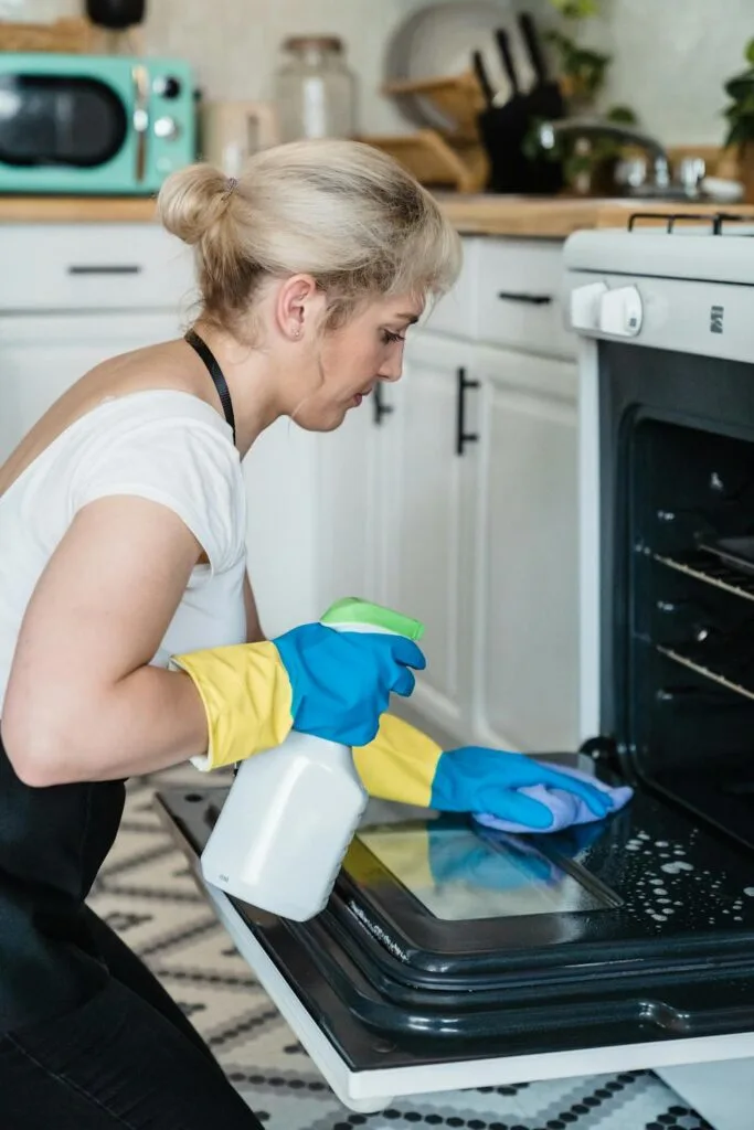 A woman wearing rubber gloves cleaning the inside of an oven with a spray bottle and cloth.