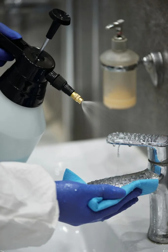 A gloved hand spraying and scrubbing a bathroom faucet to remove built-up water spots and residue.