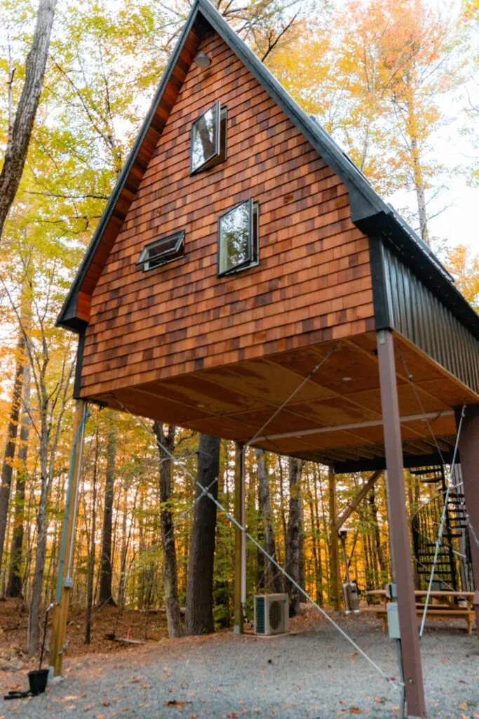 Modern A-frame cabin on stilts in a wooded setting, a unique short-term rental property surrounded by fall foliage.