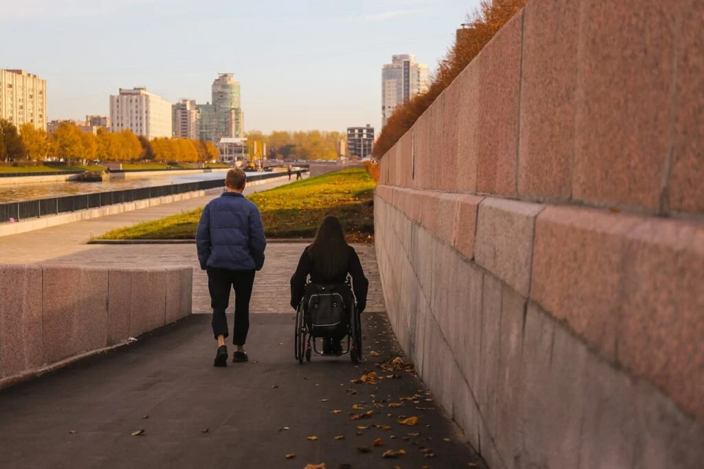 A person using a wheelchair moving along a riverside walkway beside a companion, highlighting independence, support, and NDIS participant wellbeing.