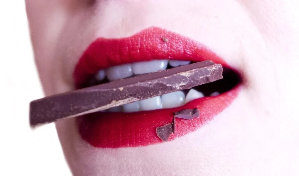 A person biting into a chocolate bar with bright red lipstick.