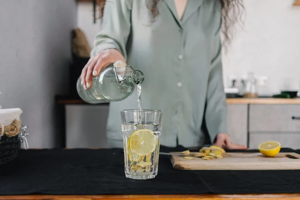Person pouring water from a glass bottle into a lemon-infused drinking glass on a kitchen counter, emphasizing hydration and simple wellness habits.