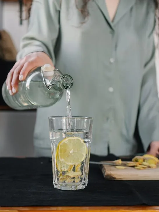 Person pouring water from a glass bottle into a lemon-infused drinking glass on a kitchen counter, emphasizing hydration and simple wellness habits.