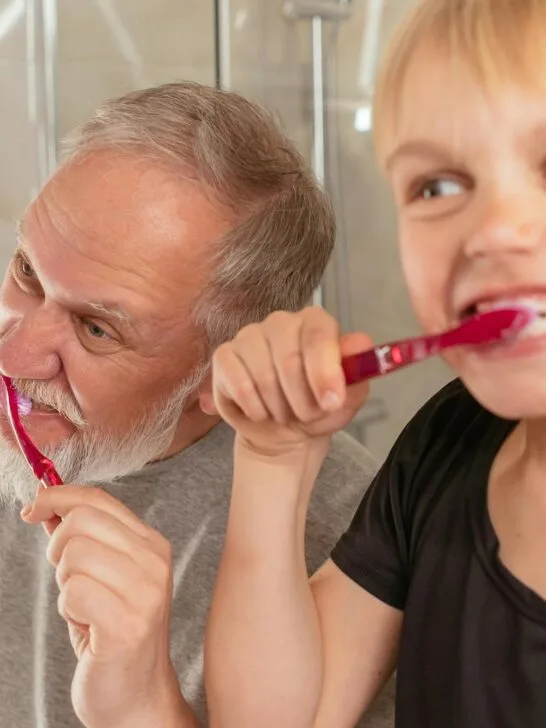 Older adult and young child brushing their teeth together in a bathroom, showing shared dental care habits across generations