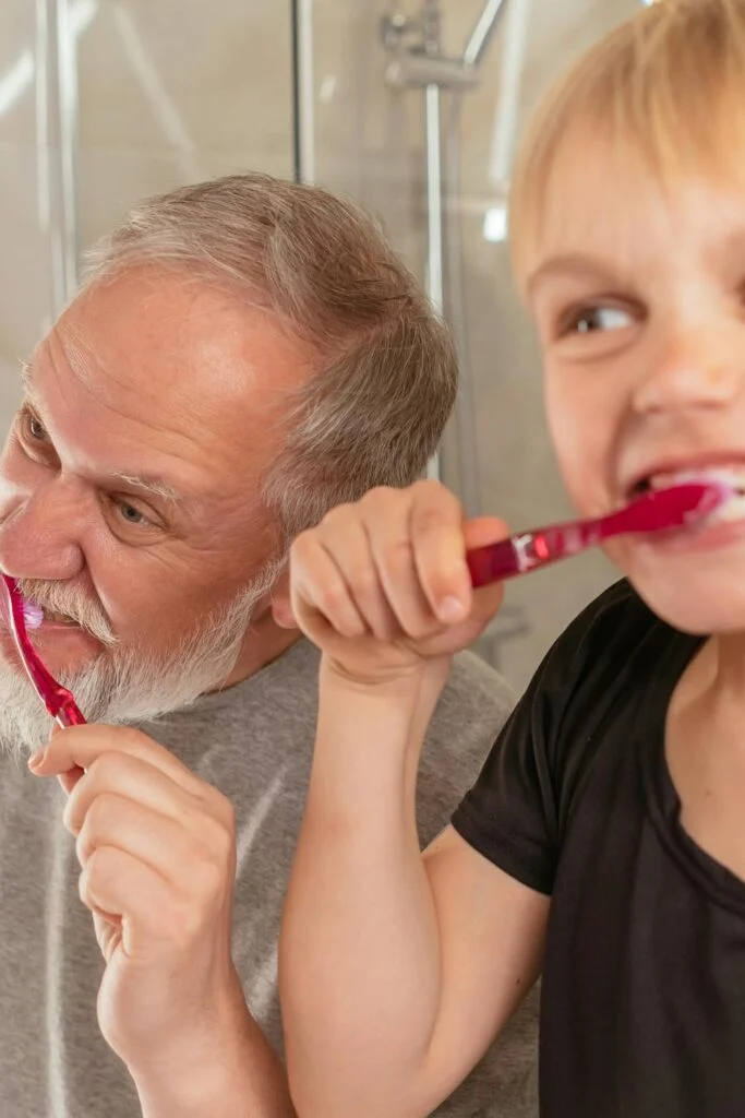 Older adult and young child brushing their teeth together in a bathroom, showing shared dental care habits across generations