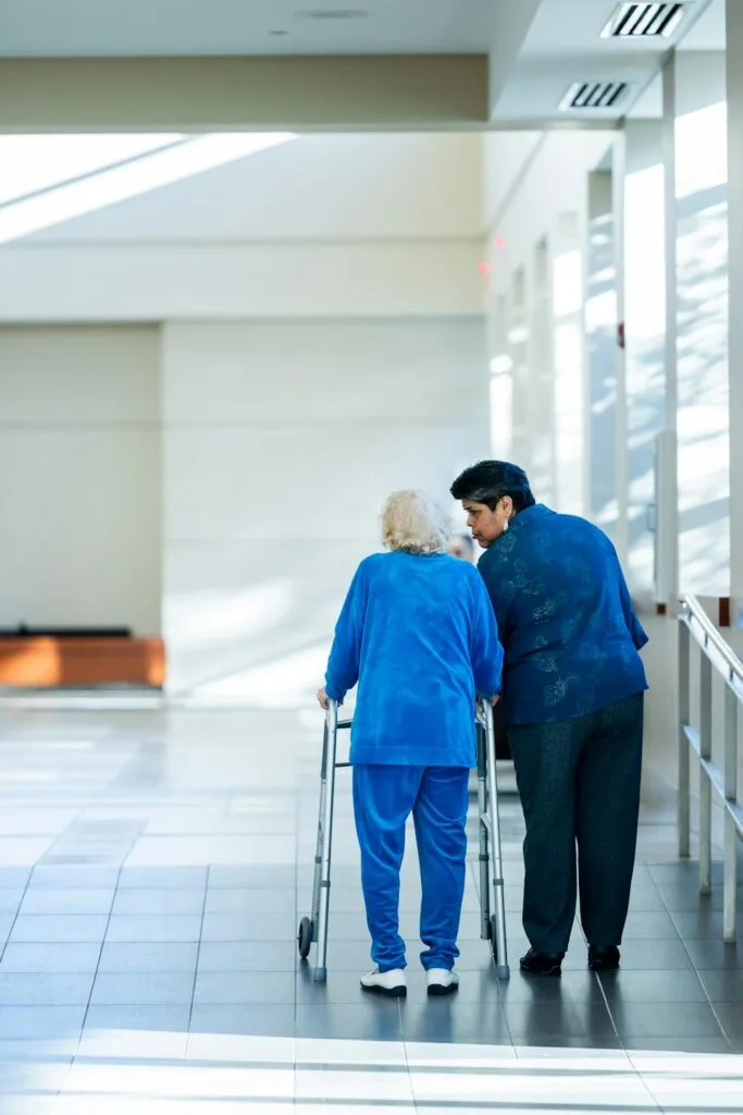 Older adult using a walker while being assisted by a caregiver in a bright indoor space, representing senior living options focused on safety and support.