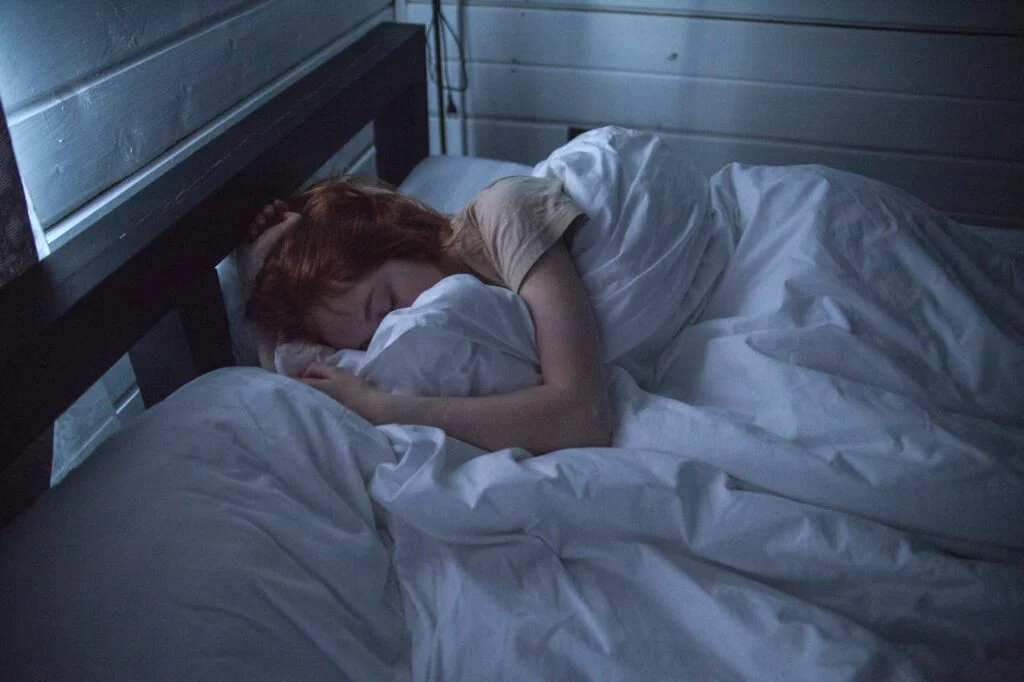 A person curled up under white blankets in a dim bedroom, resting peacefully on a soft pillow.