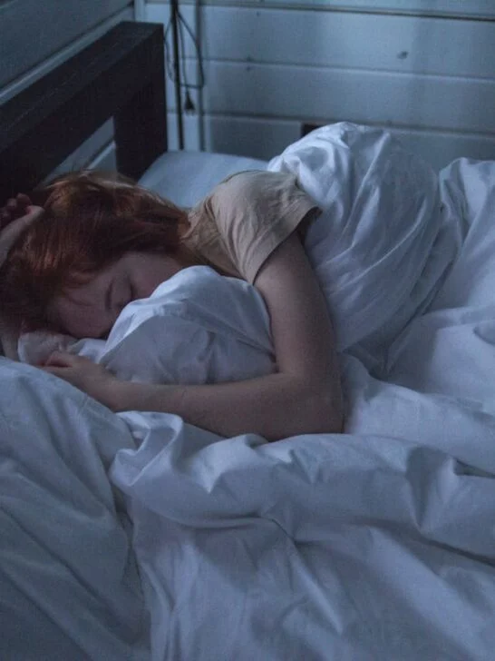 A person curled up under white blankets in a dim bedroom, resting peacefully on a soft pillow.