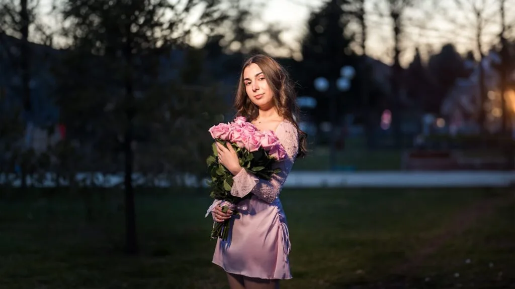 Woman standing outdoors holding a bouquet of pink roses while wearing a soft pink mini dress during early evening light.
