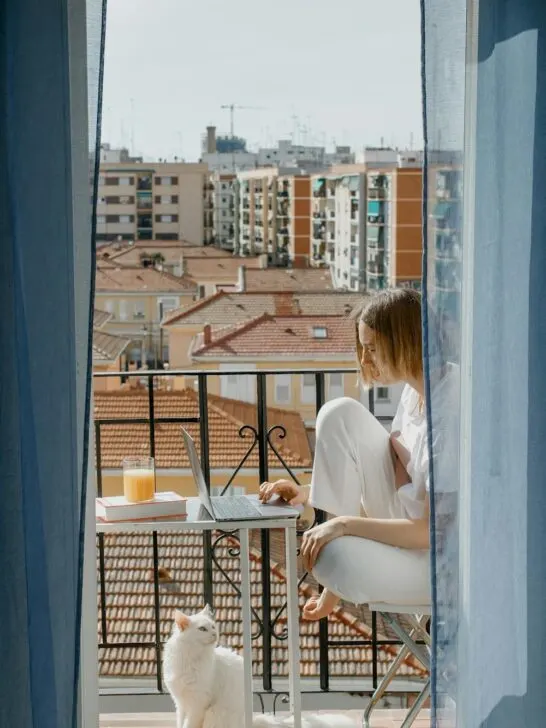 Woman working on a laptop in an apartment balcony while her cat sits nearby, representing pet approval in rentals
