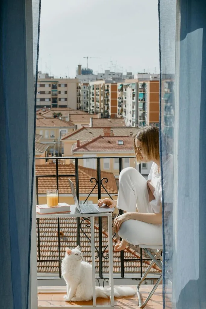 Woman working on a laptop in an apartment balcony while her cat sits nearby, representing pet approval in rentals