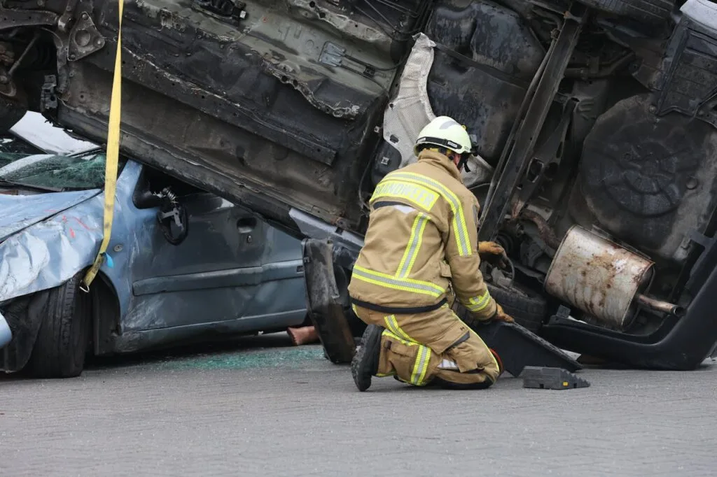 Firefighter kneeling beside overturned vehicles at a serious car crash scene while assisting trapped victims on the roadway