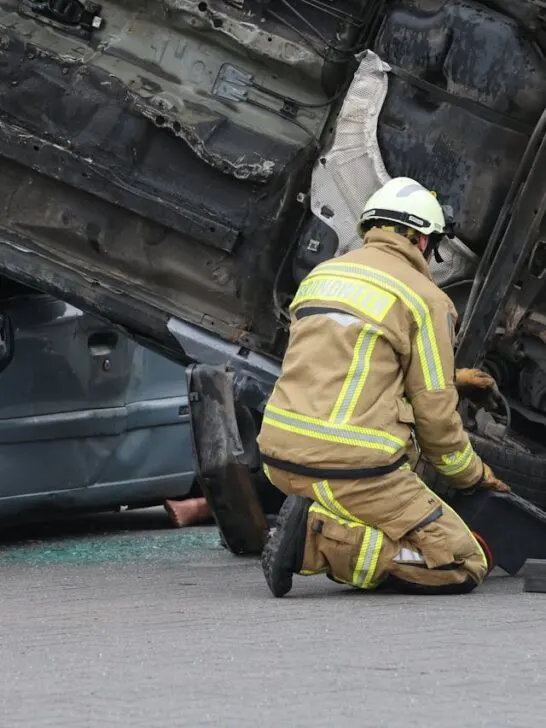 Firefighter kneeling beside overturned vehicles at a serious car crash scene while assisting trapped victims on the roadway