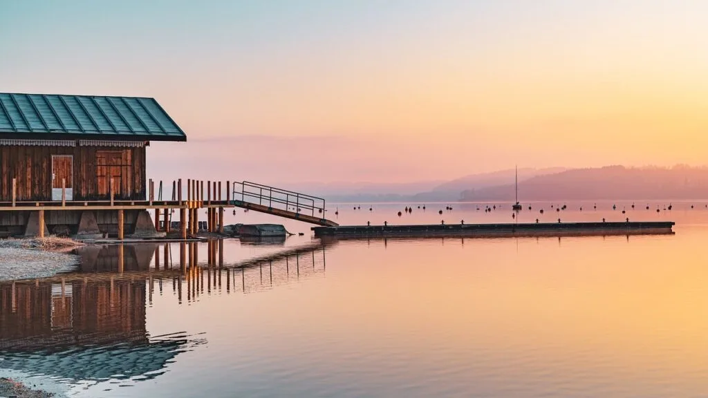 Lakeside wooden boathouse at sunset with dock and calm water reflecting golden skies, highlighting peaceful boathouse living benefits