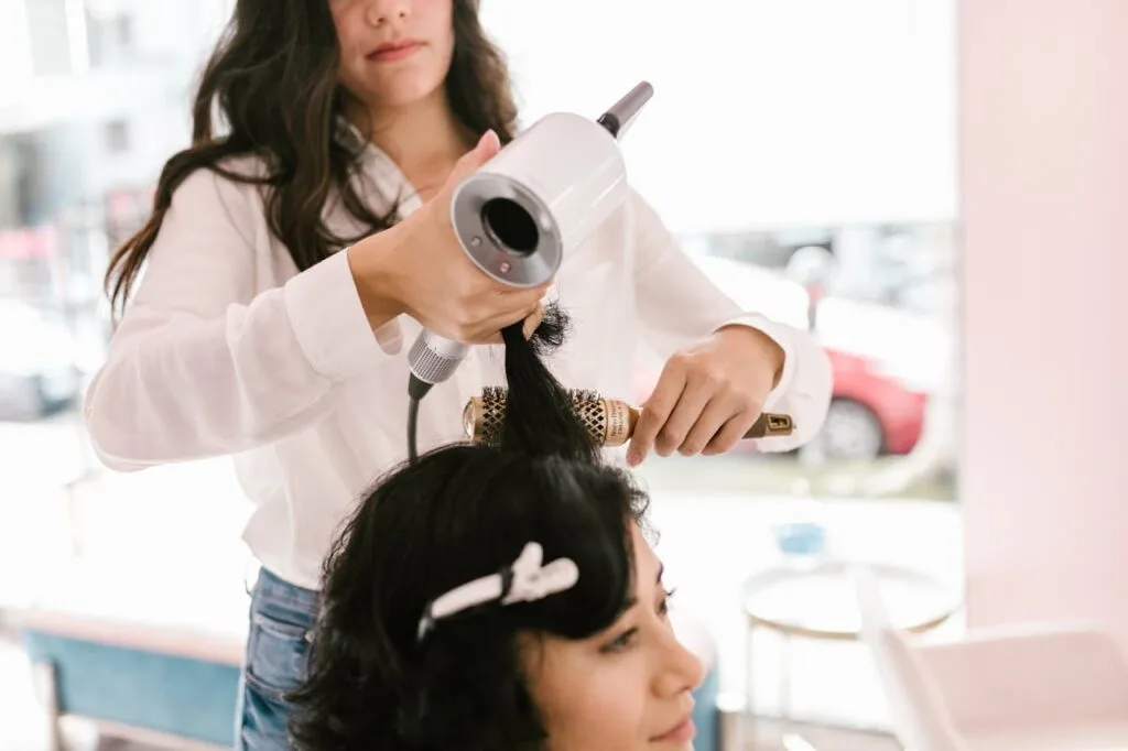 Hair stylist blow-drying a client&rsquo;s hair with a round brush inside a clean, professional beauty salon setting.