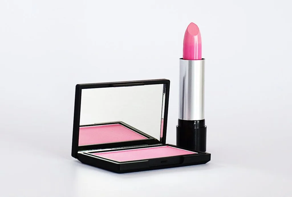 Pink lipstick in a silver tube standing beside an open blush compact with mirror on a clean white background.