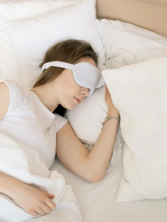 Woman resting peacefully in white bedding and eye mask, demonstrating quality bedding for better sleep