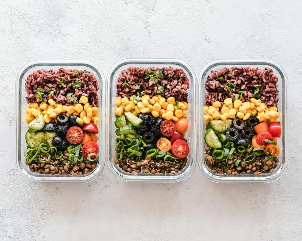 Three glass meal prep containers filled with rice, lentils, corn, cucumbers, cherry tomatoes, olives and green onions, showcasing colorful time-saving lunch ideas.