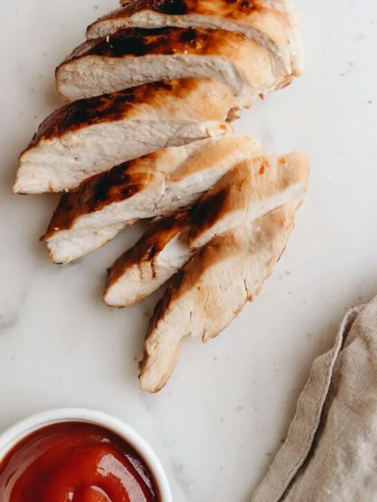Sliced grilled chicken breast arranged on a white surface with a small bowl of sauce, perfect for easy weekly meal prep.
