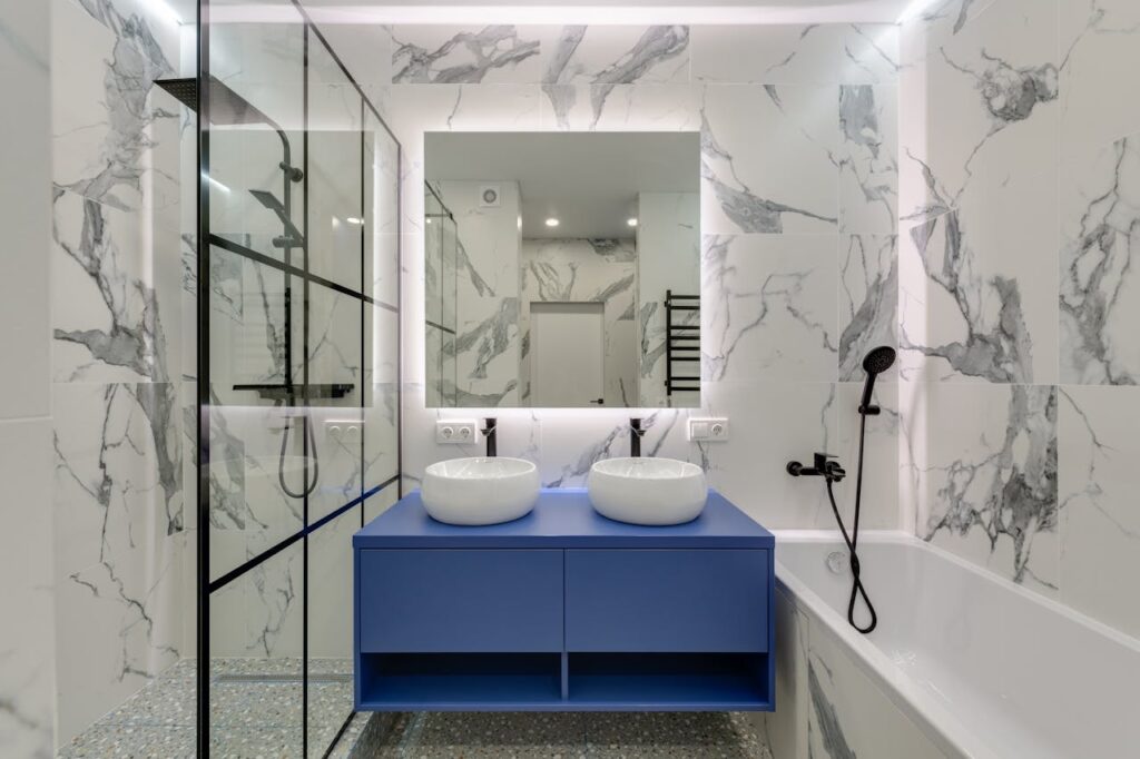 Modern luxury bathroom remodel ideas featuring marble walls, double vessel sinks, blue vanity cabinet, glass shower, and LED mirror lighting.